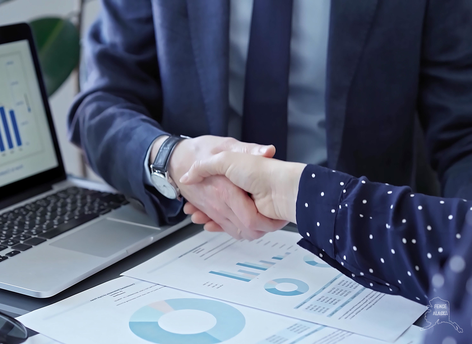 Two people in business attire are shaking hands at a desk with charts, graphs, and a laptop displaying financial data.